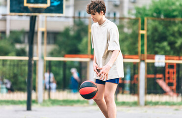 Cute teenager plays basketball at city playground. A boy holds basketball ball in his hands outside. Active life, hobby, sports for children