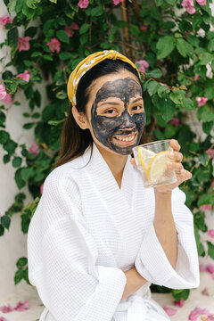 Cheerful Asian female drinking lemon water during spa procedure