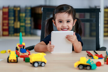 Small Asian pigtails hairstyle preschooler kindergarten girl sitting at table full of plastic wood truck toys smiling look at camera touching tablet computer in living room at home in front bookshelf