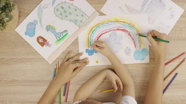 Top view seeing hands and arms of mother and her daughter kid girl are drawing and coloring beautiful rainbow pictures on white paper shows concept of parental time for happiness together activity.