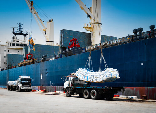Ship crane lift-off slings of sugar bags cargo from truck and load into ship hold at seaport terminal for export.