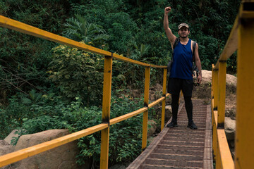Obraz na płótnie Canvas young hiker man raising his arm victorious in the middle of a bridge and carrying a red backpack surrounded by green trees in the rain forest on Cerro Ena in Costa Rica