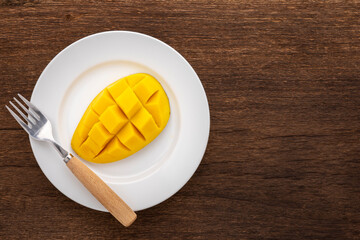 tasty fresh yellow ripe mango with fork in simply white ceramic plate on rustic natural wood texture background with copy space for text, top view