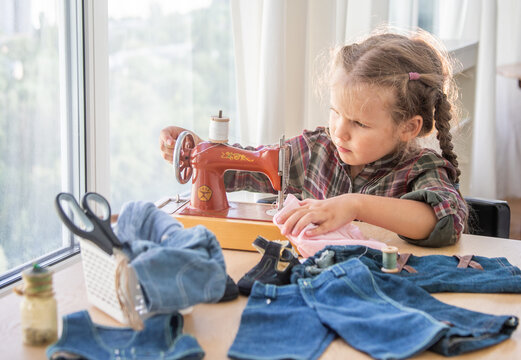 A little girl sews clothes for dolls on a toy sewing machine, a child learns to sew outfits for toys.
