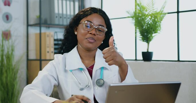 Close up of charming smiling professional cvalified black-skinned female doctor in white gawn which sitting in front of camera with computer and gesturing symbol alright