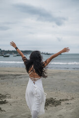 Fototapeta na wymiar Beautiful woman in white dress walking on the beach. Hands to the side. Back view. Sea background. Black long curly hair. Wind in her hair. Romantic photo.