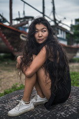 Beautiful young asian woman in black dress and white sneakers sitting on the stone near old wooden ships. Blurred background. Black long curly hair. Romantic photo.