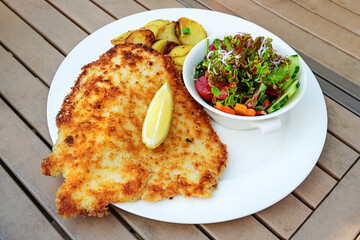 Wiener schnitzel with lemon slice, salad and fried potato on a wooden garden table in an outdoor...