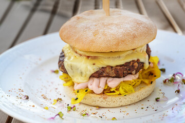 Burger with a coarse meat patty, cheese, mayonnaise and saffron cabbage salad, garnished with spices and radish sprouts on a white plate and a wooden table, copy space, selected focus