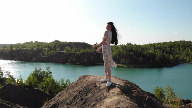 beautiful slender woman in long white dress approaches edge of abyss and spreads her arms to sides, she conquered the top, enjoys view. lady stands on mountain with blue lake in the background.