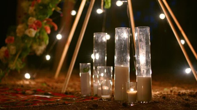 Candles burning in glass vases, flasks on carpet near bohemian tipi arch decorated with roses and flowers wrapped in fairy lights on outdoor wedding ceremony venue in pine forest. Boho rustic decor.