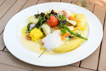 Vegetables with fried fish and potatoes in saffron sauce, garnished with herbs and foam on a white plate and a wooden table, selected focus