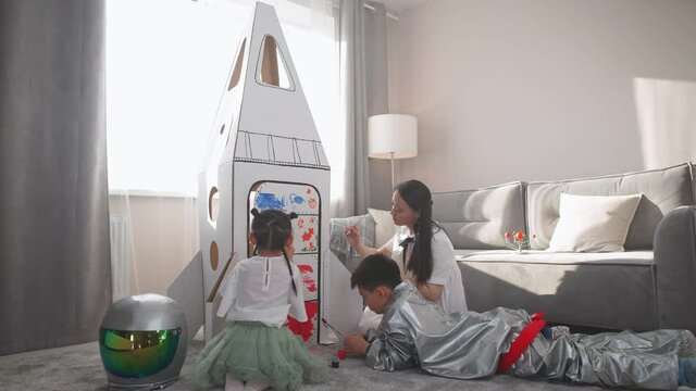 Asian female with her kids play in the living room at home, a boy in an astronaut costume lying on the floor, children together with their mother paint on a cardboard model of a spaceship with paints.