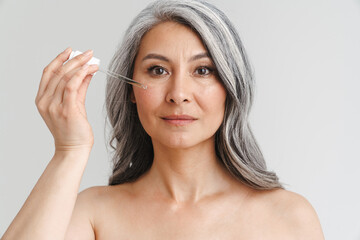 Mature shirtless woman with grey hair applying face serum