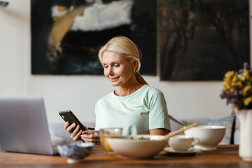 Blonde mature woman using laptop and cellphone while having lunch