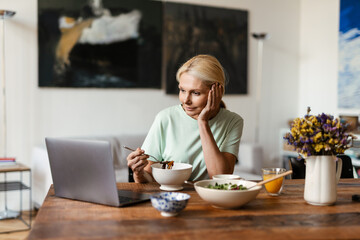 Blonde mature woman using laptop while having lunch at home