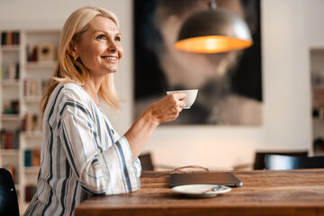 Blonde mature woman smiling and drinking tea while sitting at table