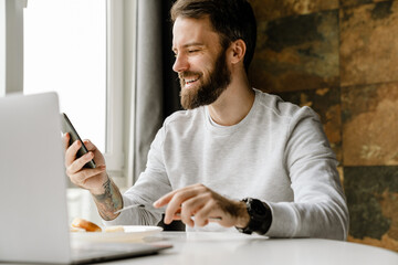 Fototapeta na wymiar Bearded man smiling and using cellphone while working with laptop
