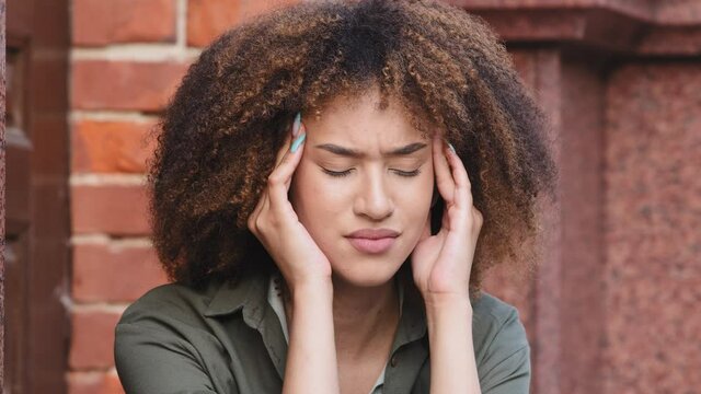 Face of unhappy young African American woman siting outdoors rubs temple, closed eyes suffers headache painful feelings chronic migraine hurt. Break up, difficult period of life concept closeup view