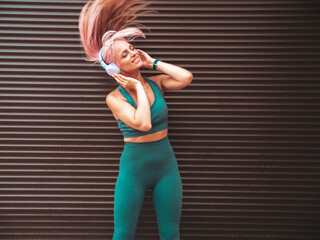 Fitness smiling woman in green sports clothing with pink hair. Young beautiful model with perfect body.Female in the street near roller shutter wall.Listening music in wireless headphones.Shakes hair
