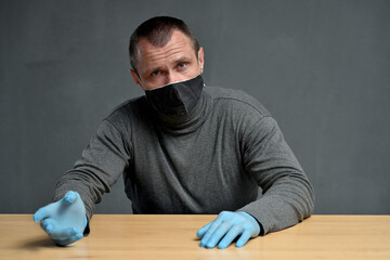 Fototapeta premium Concept on a gray background an adult man in a mask and blue gloves sits at a table and looks at the camera while talking with the interlocutor