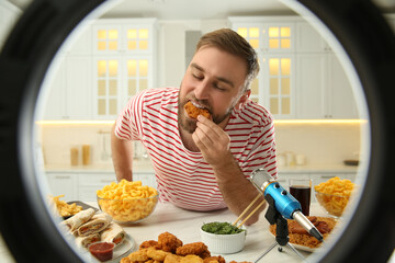 Food blogger eating near microphone at table in kitchen, view through ring light. Mukbang vlog