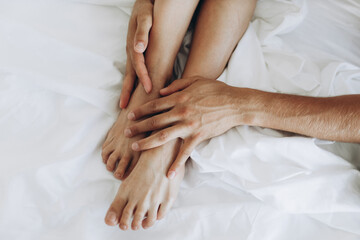 Fototapeta na wymiar Thin smooth tender legs hug the hands of a man on white sheets. 