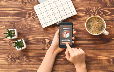 Woman holding mobile phone with open page of delivery service application on wooden table