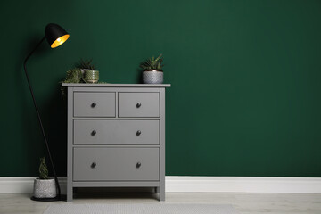 Modern chest of drawers with houseplants and lamp near green wall indoors. Space for text