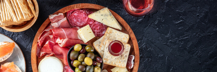 Charcuterie and cheese board panorama with red wine and olives, shot from the top on a black background with copy space. Spanish tapas or Italian antipasti