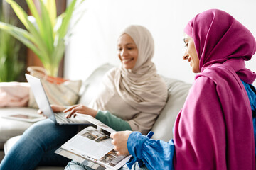 Two women reading magazine and using laptop while sitting on sofa