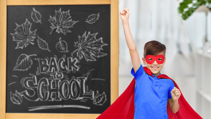 education, learning and people concept - happy boy in red super hero cape and mask showing fists over chalkboard with back to school lettering on background