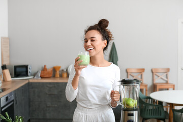 Young African-American woman drinking fresh smoothie in kitchen