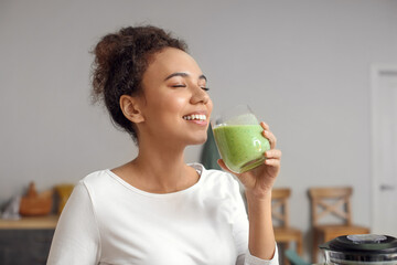 Young African-American woman drinking fresh smoothie in kitchen