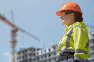 An engrossed female engineer works against the background of a multi-storey building under construction. Portrait of a young architect of a construction site, protective equipment. Selective focus.