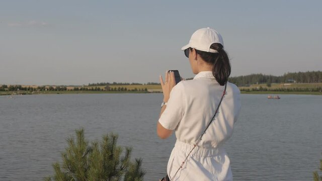 Young woman in white baseball cap taking a picture of the lake at sunset.