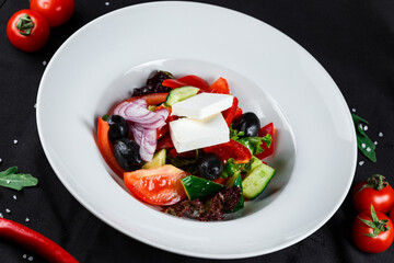 greek salad with olives, bell peppers, onions and feta cheese in a white plate on a dark background with ingredients close-up top view