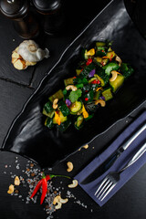 chinese salad of crushed pickled cucumbers with cashews and herbs in a black curved plate on a dark decorated background.