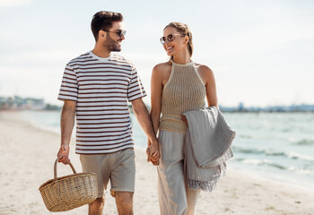 leisure, relationships and people concept - happy couple with picnic basket and blanket walking along beach