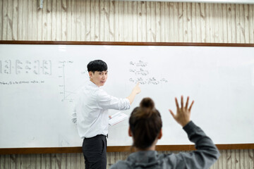 An Asian male teacher in a classroom near the whiteboard is writing and teaching a lesson with a...