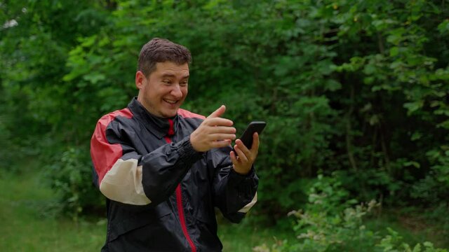 Excited happy man looking at smartphone in forest. Young adult 30s person with positive emotions smile about good news. Male watch on phone screen and joy fine information or win in online lottery