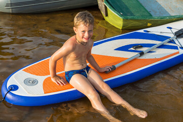 a cute boy in swimming trunks is sitting on a sup board in the water near the shore