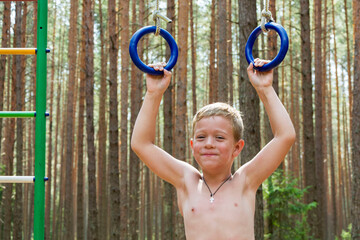 Portrait of a cute boy pulling himself up on gymnastic rings in nature against the background of a...