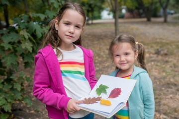 Two adorable friends girls collecting colorful leaves for herbarium on warm autumn day in the forest. Children exploring nature outside. Kids in fall concept