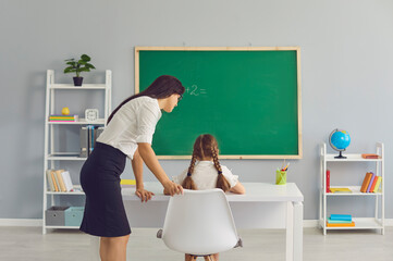 Female teacher helping little girl on math lesson at classroom, space for text. Tutor and pupil working together during class at school