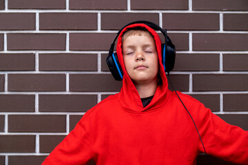 Photo of concentrated boy child with eyes closed listening music with headphones. Freedom of music. Teenager boy gets pleasure from music.Boy in red clothes.