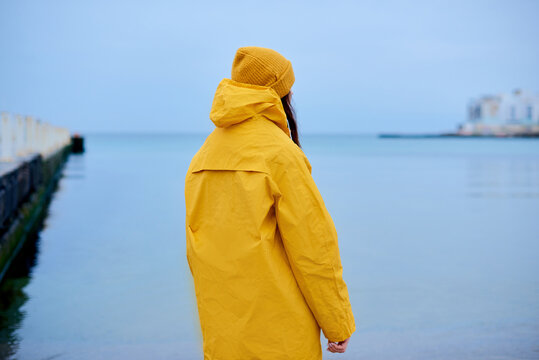 sadness woman standing alone on beach and look at distant city. loneliness woman in yellow rain coat walking coast