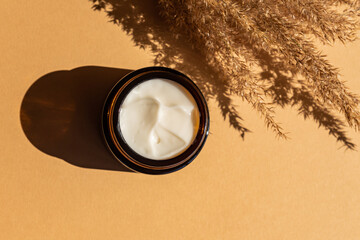 Moisturizer cream jar in amber glass bottle and dry flowers reeds on beige background. Flat lay,...