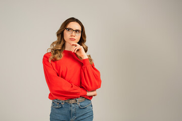 pensive and curly woman in eyeglasses looking away isolated on grey