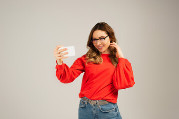 positive woman in eyeglasses taking selfie on smartphone isolated on grey